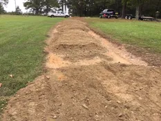 Wide dirt trench dug across a green lawn with soil mounds along the sides, extending toward the background.